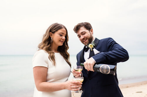 Groom pours champagne for bride on beach after their destination wedding ceremony in Union Pier, Michigan
