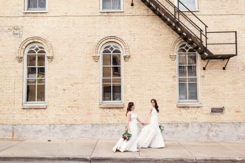 Two brides holding hands walk in front of cream city brick building after Mercantile Hall wedding