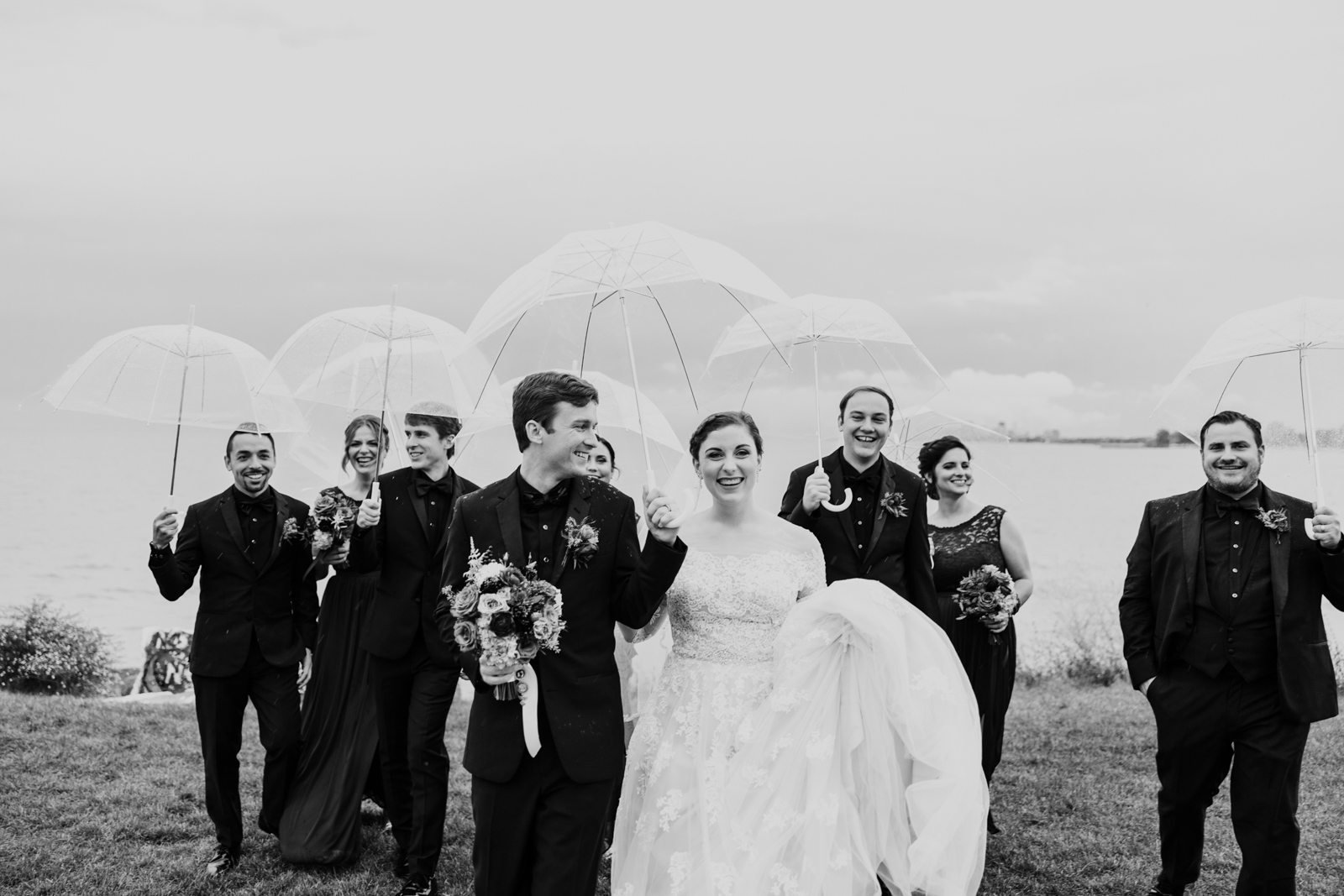 Wedding party walks along lakefront with umbrellas on rainy day by documentary wedding photographer Emma Mullins Photography