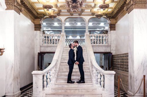 Two grooms kiss on marble staircase before their Chicago Athletic Association Hotel wedding at Cindy's Rooftop