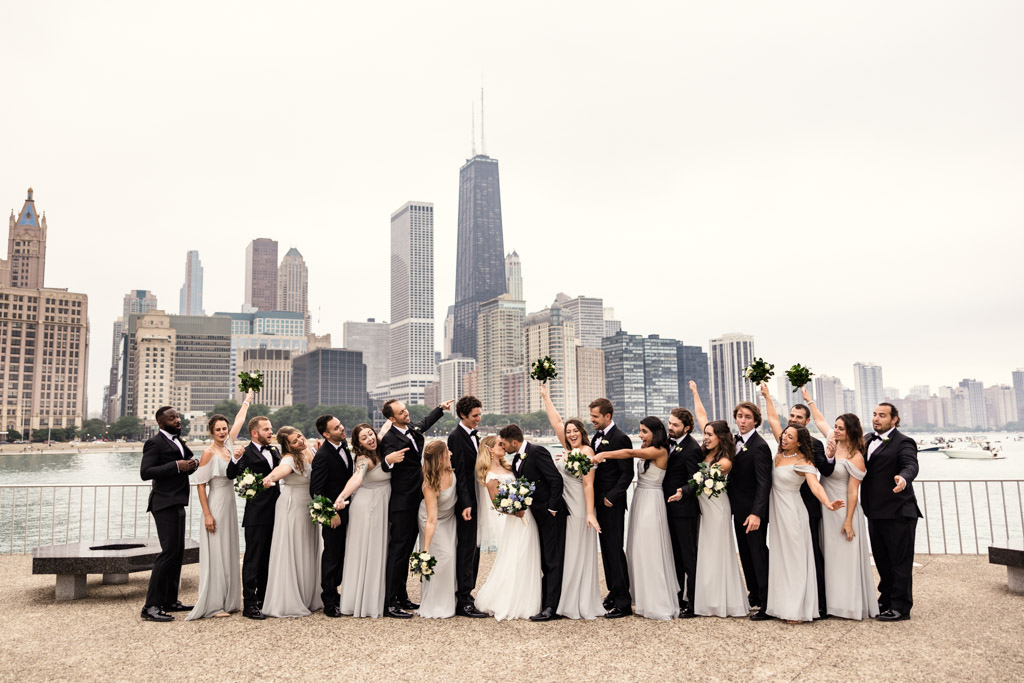 Wedding party cheers while bride and groom kiss with Chicago skyline in background at Olive Park