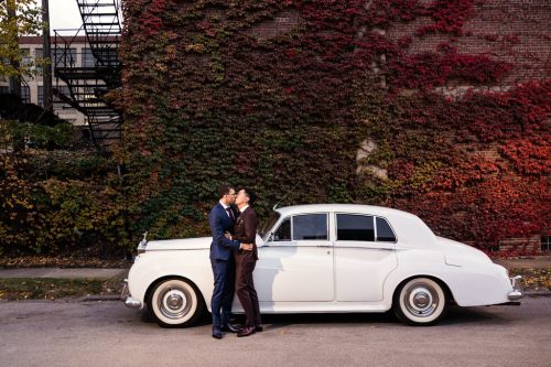 Groom and groom kiss in front of Rolls Royce with fall foliage at their Ovation Chicago wedding