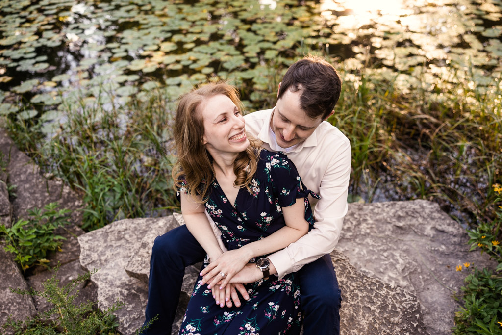 Couple smiles at each other while sitting on rock at Alfred Caldwell Lily Pool engagement session in Chicago