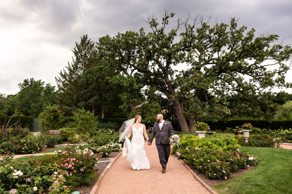Bride and groom walk in rose garden with large Oak tree before their summer Cantigny wedding ceremony in Wheaton, Illinois