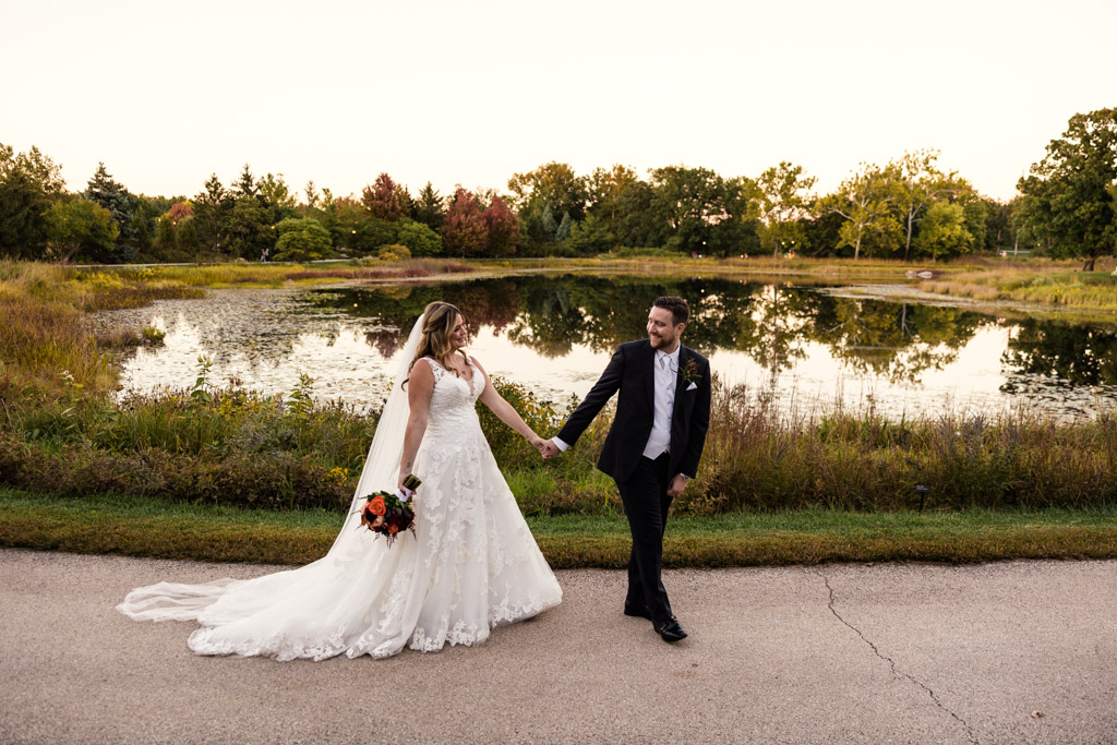 Bride and groom walk along Meadow Lake at sunset during their fall Morton Arboretum wedding