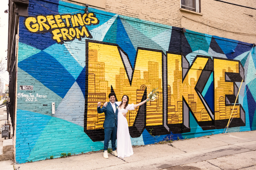 Bride and groom cheer in front of Greetings From MKE mural on their spring Milwaukee elopement day