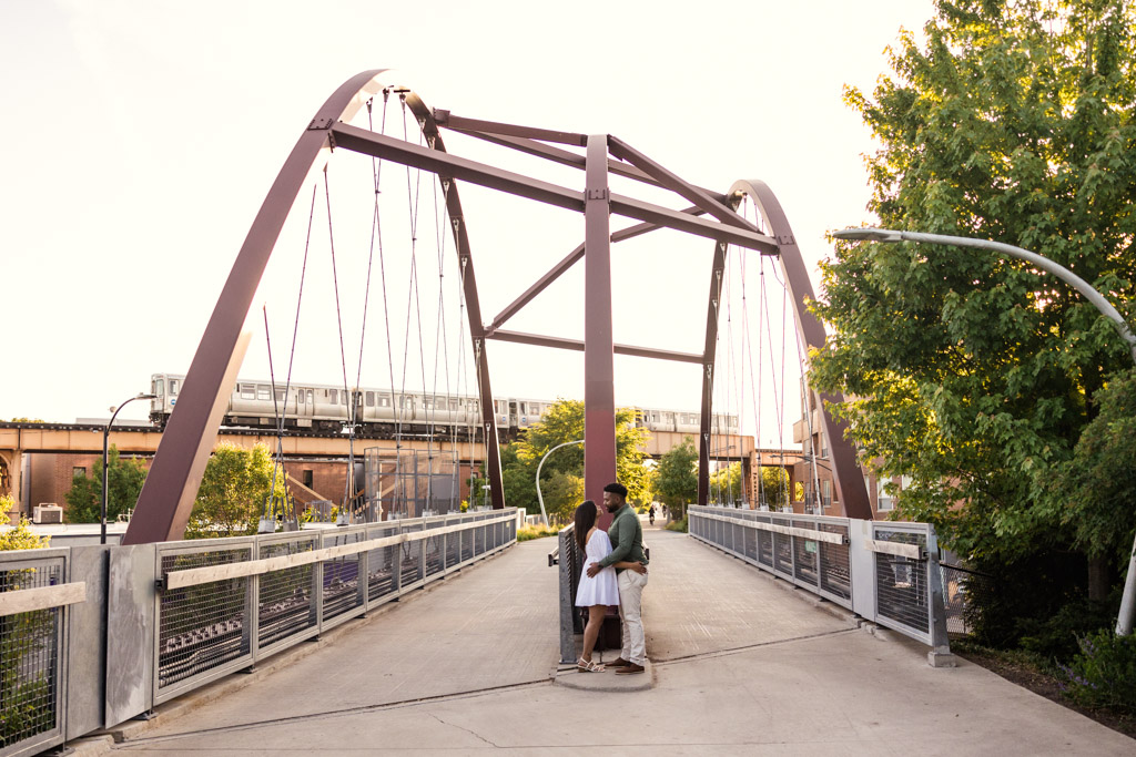 A couple embraces on a modern pedestrian bridge during their Wicker Park engagement, with a train passing by in the background.