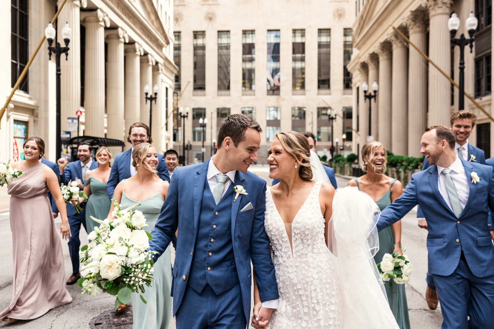 Happy bride and groom walk down LaSalle Street downtown Chicago in front of Board of Trade building with their wedding party by documentary wedding photographer Emma Mullins Photography