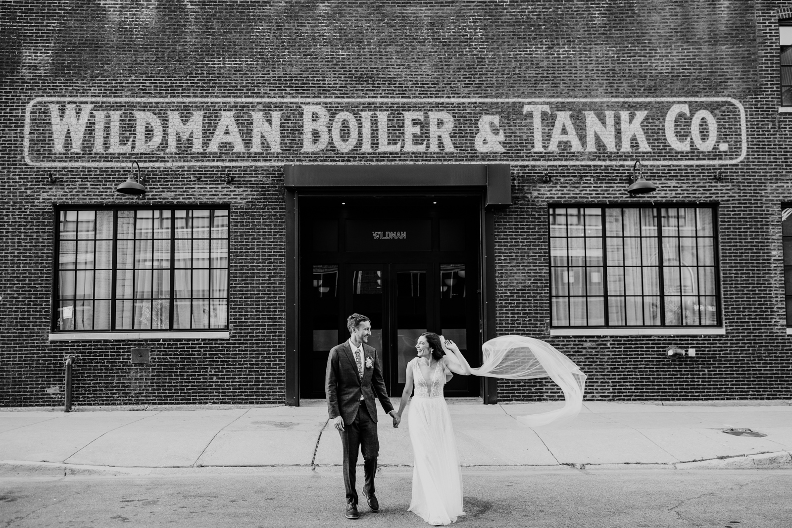 Black and white photo of bride and groom walking outside Wildman BT wedding venue with flowing veil by Chicago wedding photographer Emma Mullins Photography