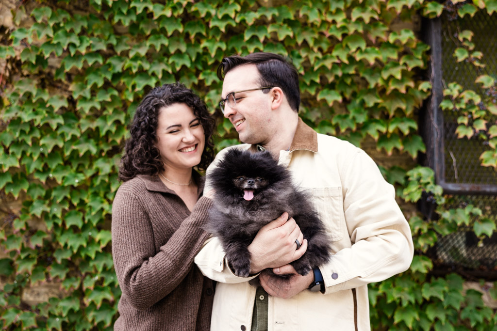 Couple holds Pomeranian while smiling in front of ivy wall during their Ravenswood neighborhood engagement session