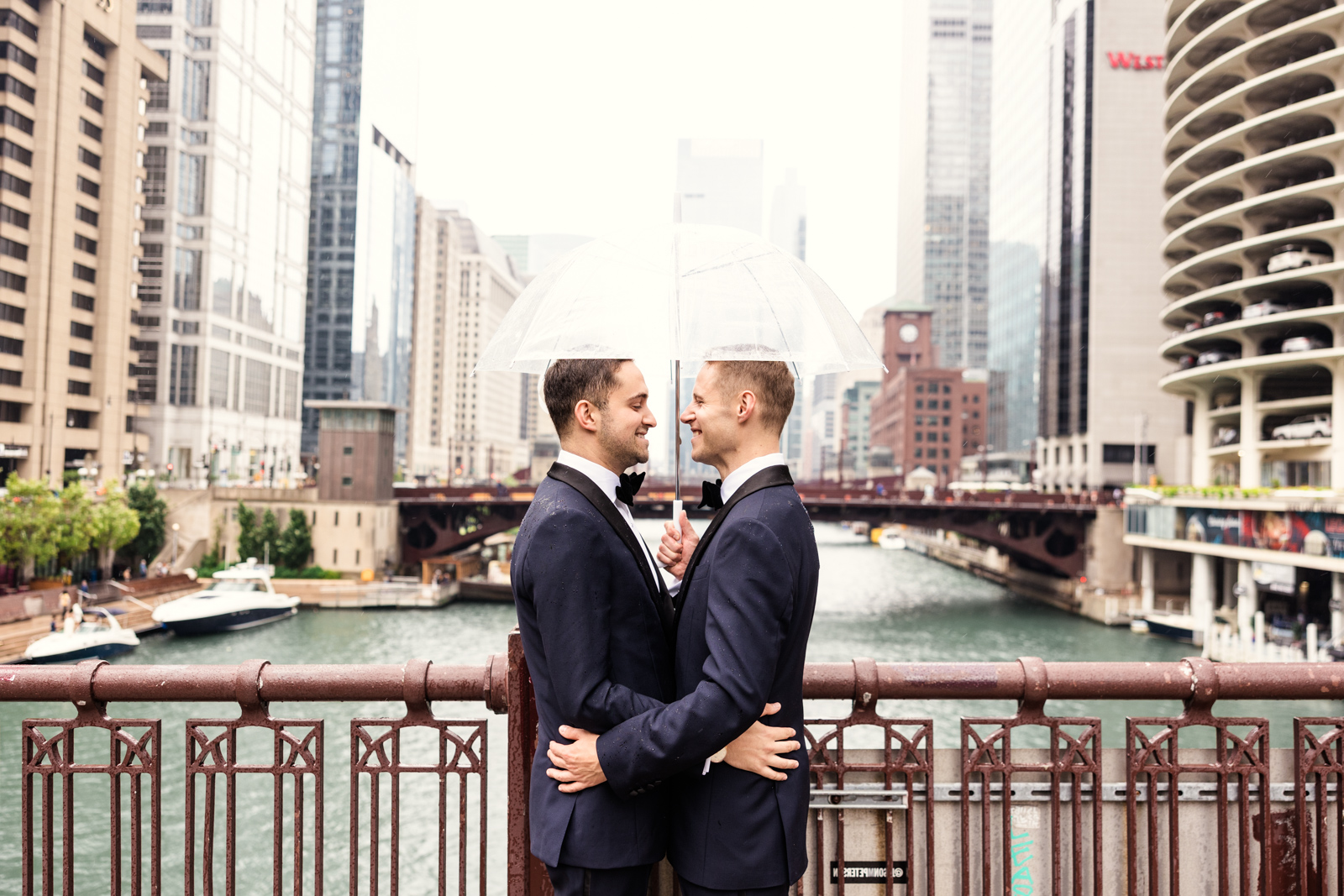 Two grooms stand on bridge under umbrella overlooking Chicago River by Emma Mullins Photography