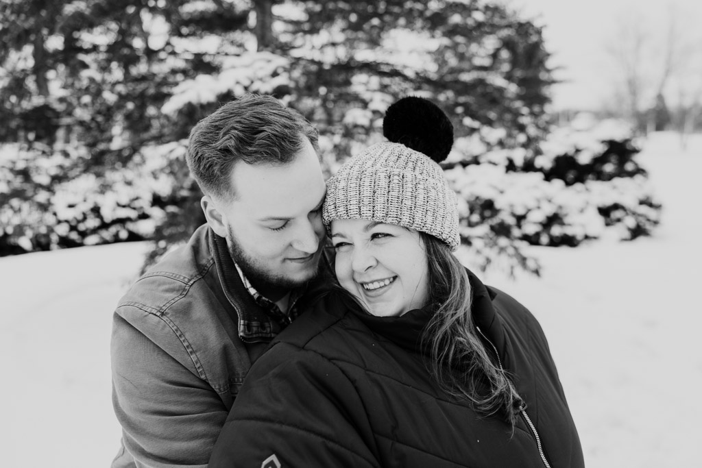 Candid photo of happy couple in coats and hats keeping each other warm during snowy winter engagement session