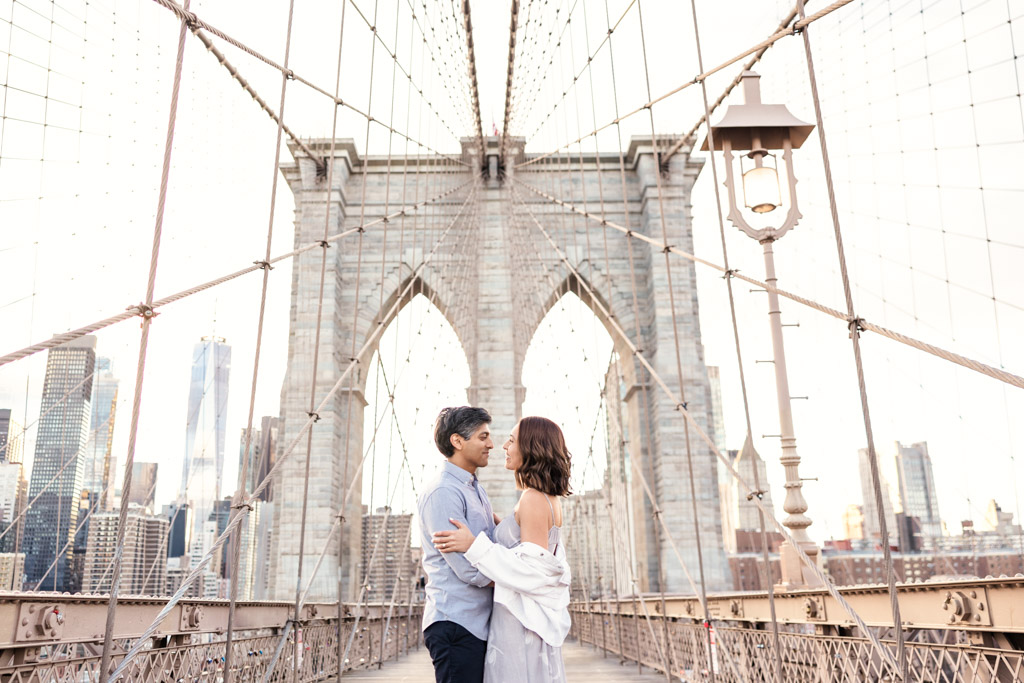 Creative Brooklyn Bridge engagement photo of couple standing on bridge with New York City skyline at sunset