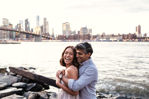 Couple laughs standing on the bank of East River with Manhattan skyline and Brooklyn Bridge in background during romantic spring engagement session