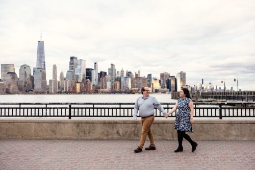Engaged couple walks along waterfront at Liberty State Park in New Jersey with Manhattan skyline view