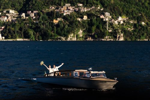 Just married bride and groom cheer while riding wooden boat on Lake Como after Villa Pizzo wedding ceremony