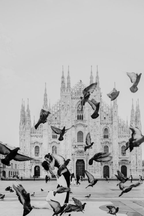 Creative wedding photo of bride and groom kissing in front of Duomo di Milano with pigeons flying around them
