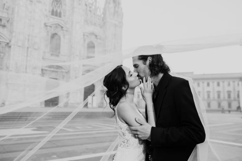 Romantic downtown Milan wedding photo of bride and groom kissing under flowing veil in front of Duomo at sunrise