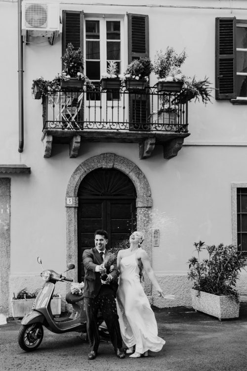Just married bride and groom pop a bottle of Prosecco on Italy street with Vespa after Lake Como elopement