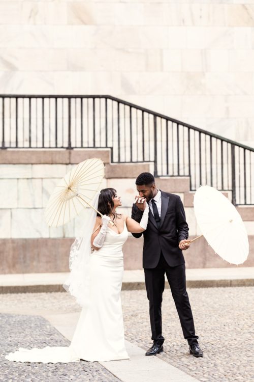 Downtown Milan wedding photo of groom kissing bride's hand while holding parasols in front of Museo 900