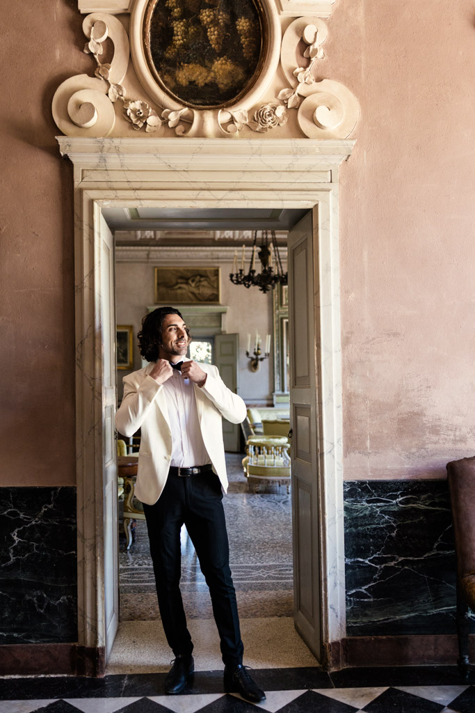 Groom in white tuxedo jacket adjusts bow tie at historic Villa Pizzo wedding venue in Lake Como, Italy
