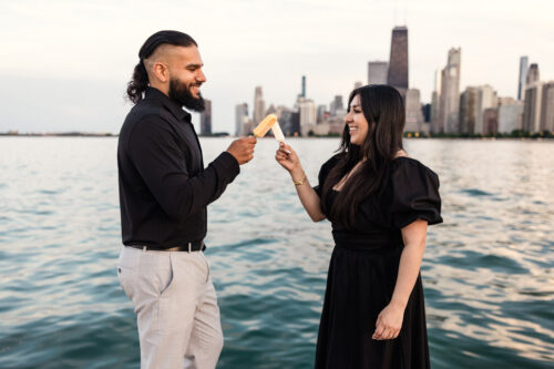 Fun photo of couples cheersing their paletas in front of Chicago skyline during their summer lakefront engagement session