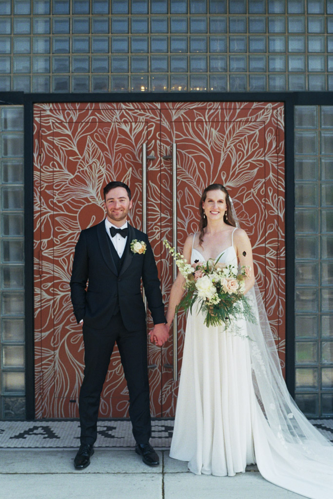 Bride and groom hold hands in doorway of The Arbory Chicago wedding venue in front of coral pink floral mural