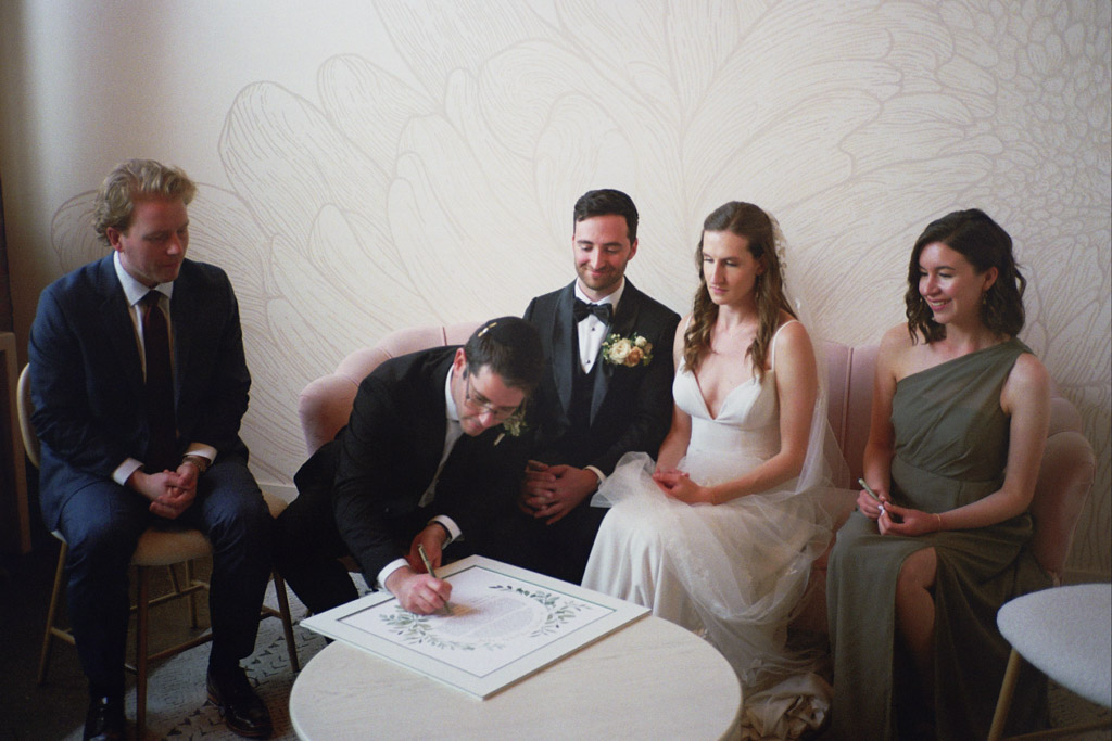 Bride and groom watch on velvet couch as witness signs their Ketubah in bridal suite at The Arbory Chicago