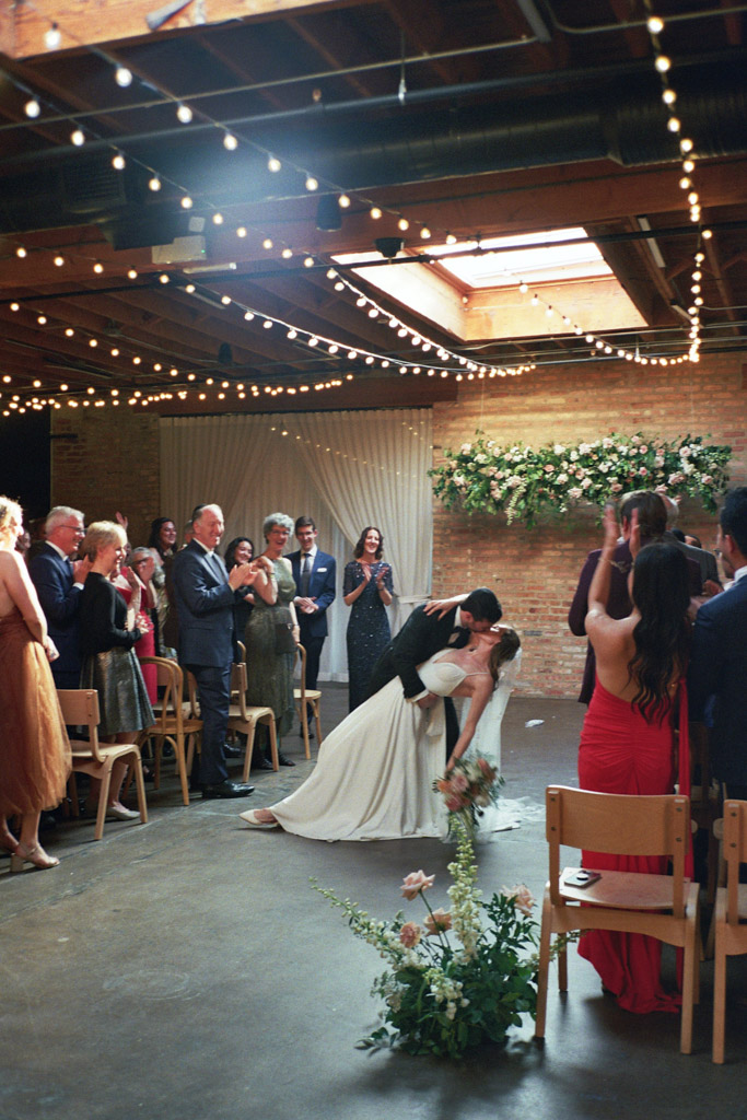 Bride and groom dip and kiss mid-aisle while guests applaud after their Arbory Chicago wedding ceremony captured on film