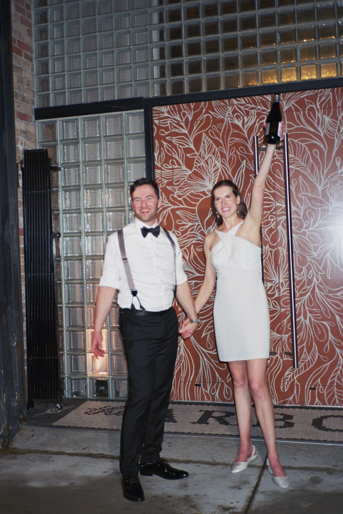 Bride and groom celebrate with bottle of sparkling wine in front of colorful door mural at The Arbory Chicago