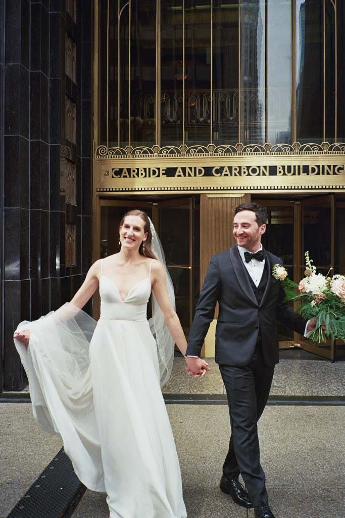 Happy bride and groom hold hands while walking outside the Carbide and Carbon building in Chicago captured on film