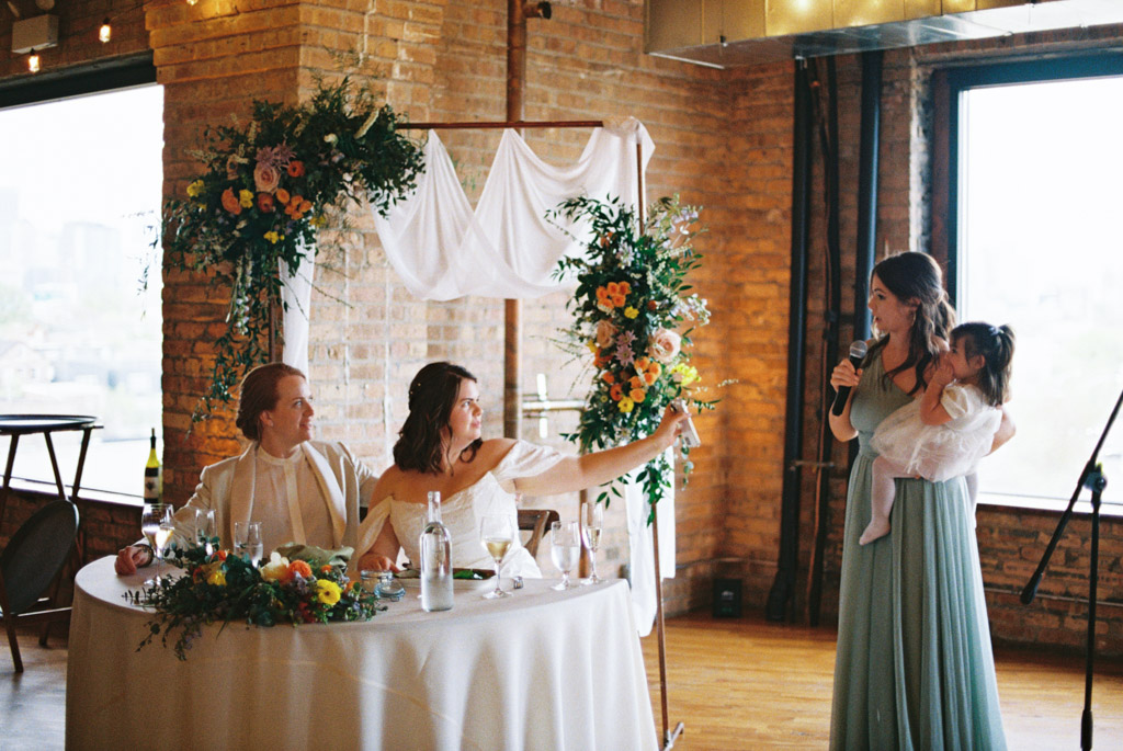 Bride's sister gives speech while holding toddler and brides sit at head table holding up phone for speaker to read from at Lacuna Lofts wedding reception