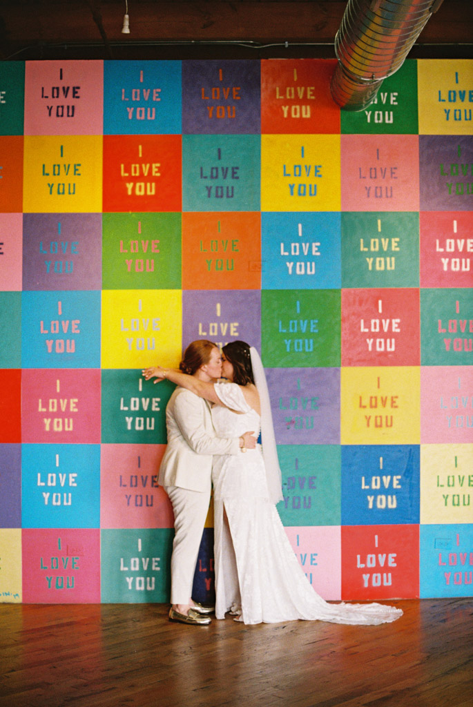 Bride and bride kiss in front of colorful I Love You wall at Lacuna Lofts wedding venue