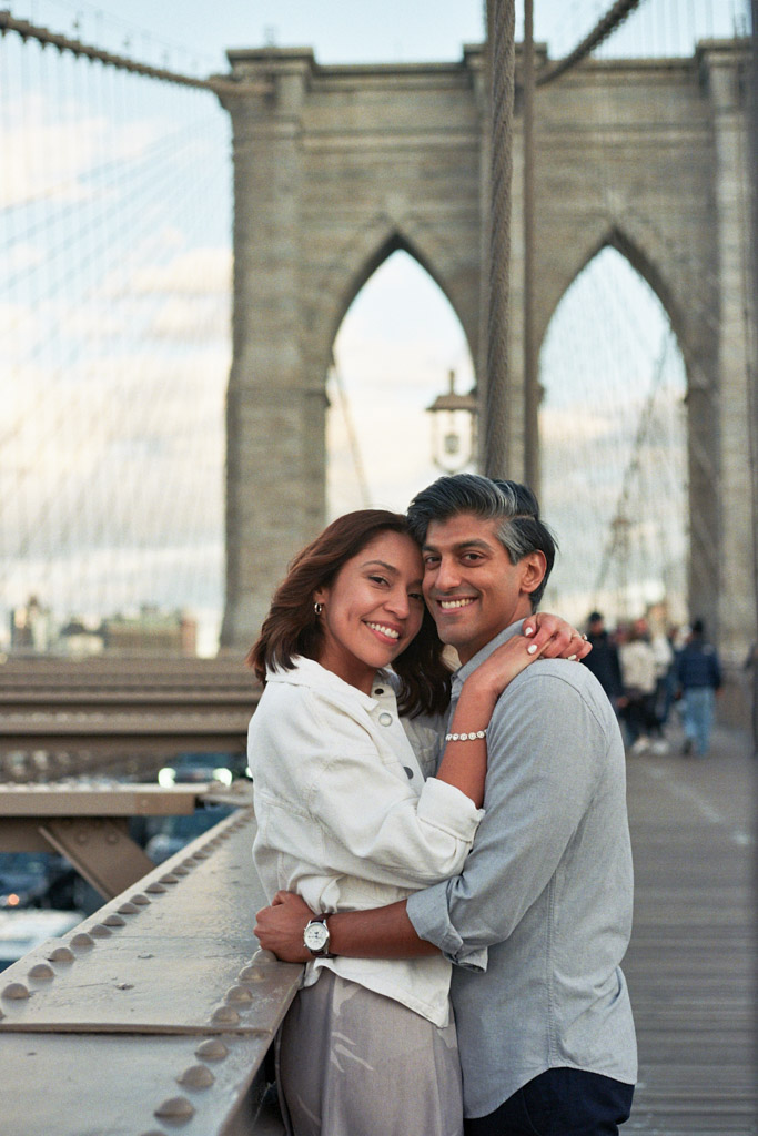 Happy couple smiles while standing on Brooklyn Bridge during their spring New York City engagement session