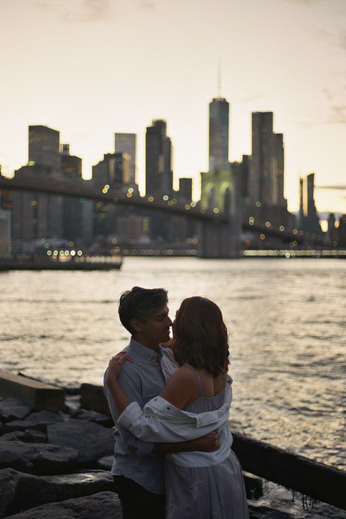 Romantic silhouette of couple standing on rocks during DUMBO engagement session with Manhattan skyline and Brooklyn Bridge in background