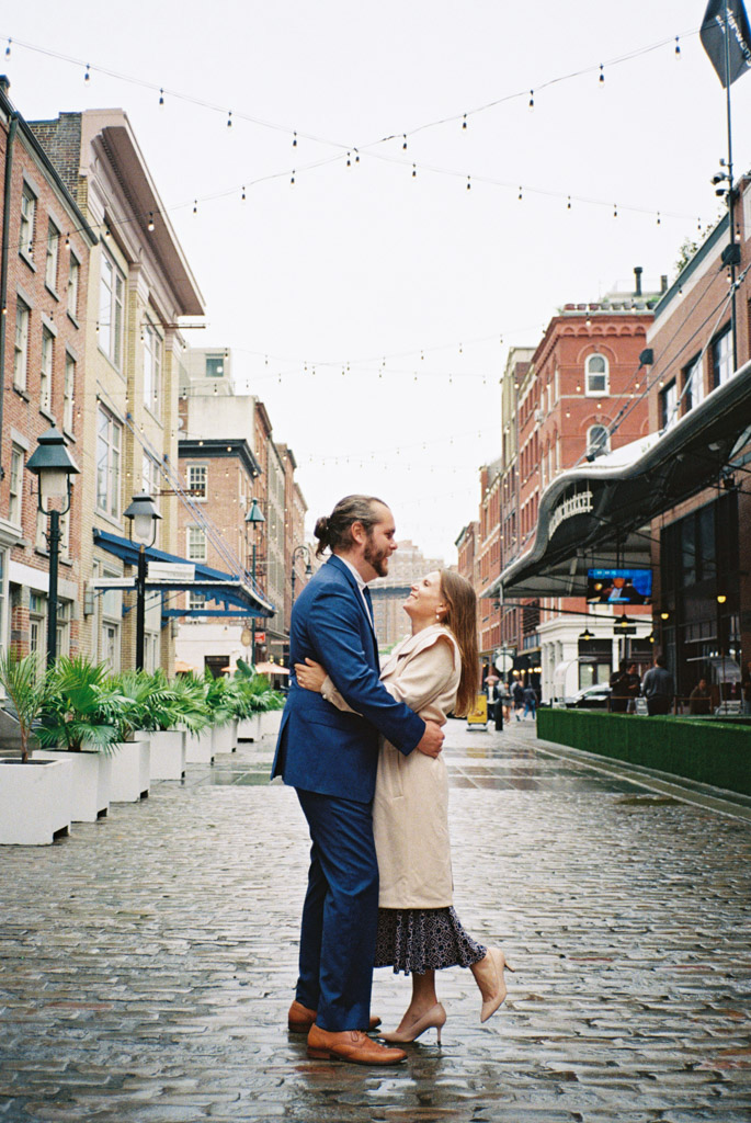 Couple stands in cobblestone street at South Street Seaport in the rain during their spring New York City engagement session