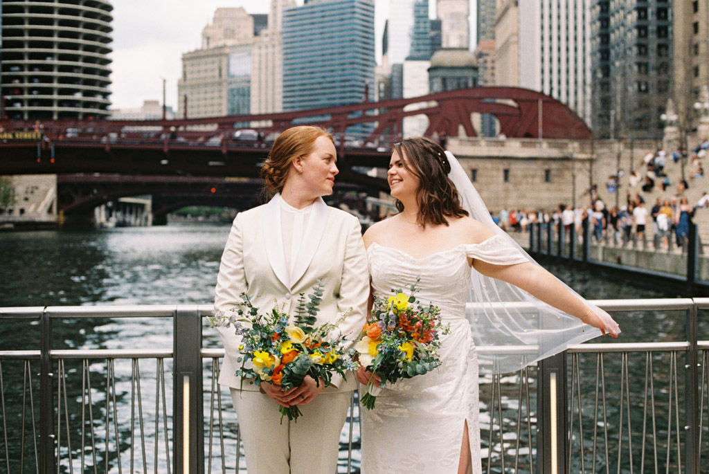 Bride and bride stand with colorful florals on Chicago bridge after wedding photos at The Rookery Building