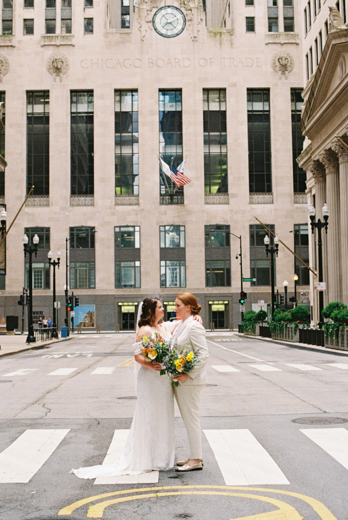Bride and bride stand in downtown Chicago street in front of Board of Trade Building with spring floral bouquets