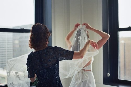 Film photo of bride adjusting her veil with the help of another woman in a dark dress, standing in front of large windows at the Pendry Chicago hotel