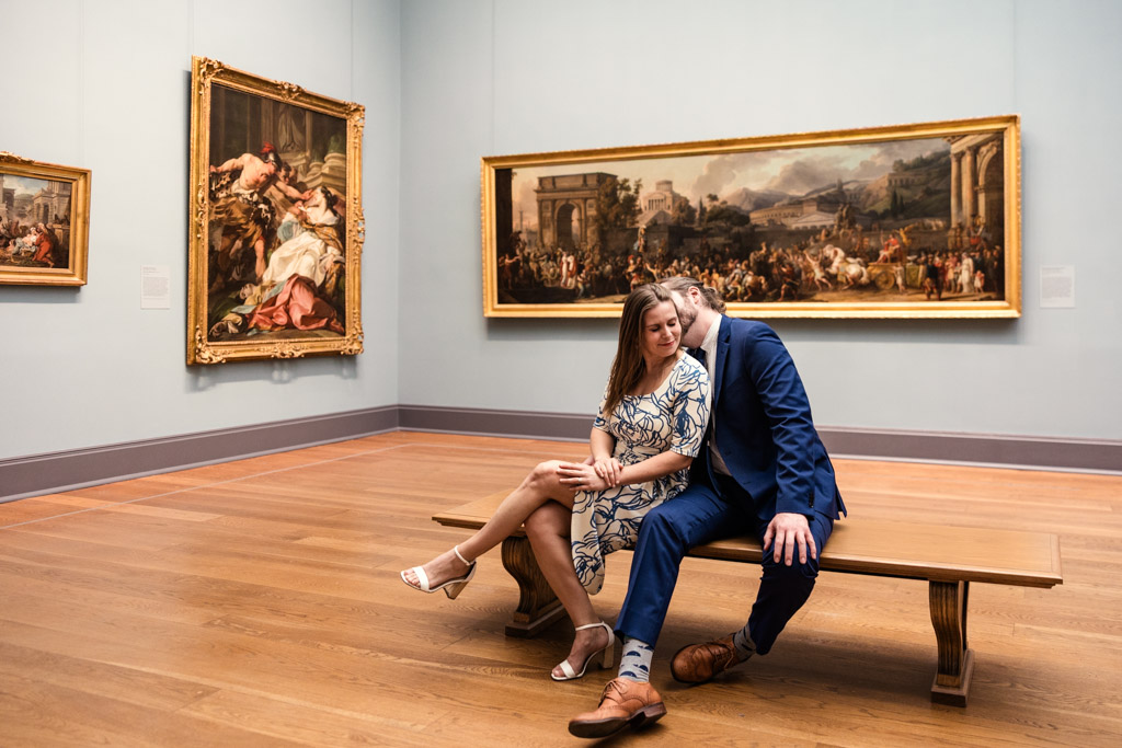 A couple sits closely on a bench in an art museum, surrounded by large framed paintings, capturing a quiet moment of their New York City engagement.