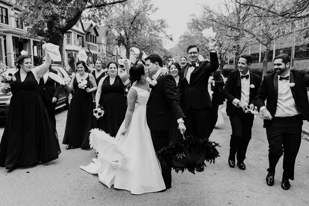 Bride and groom kiss while holding umbrellas and guests parade behind them during New Orleans wedding Second Line