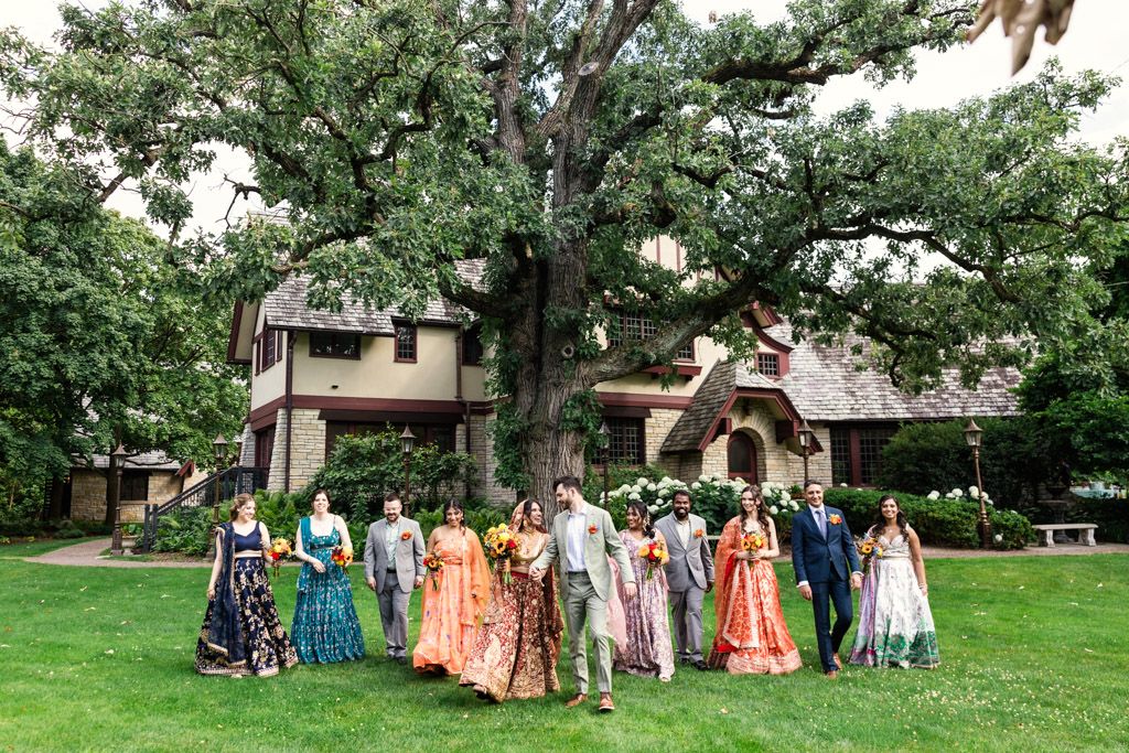 Candid photo of wedding party walking in colorful Indian attire and Western attire in front of oak tree at The Redfield Estate wedding property in Glenview, Illinois