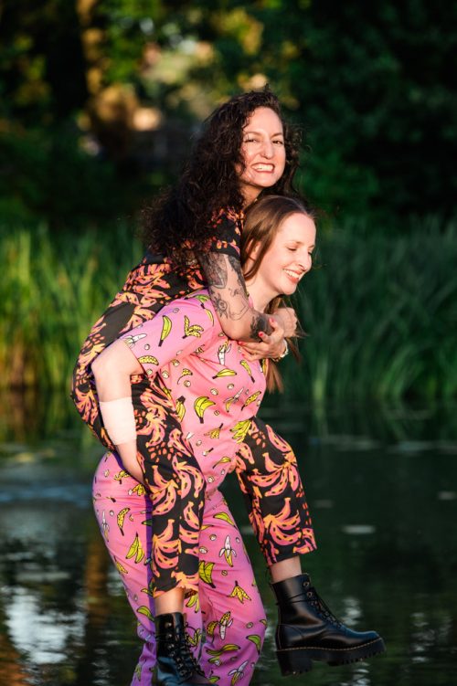Candid photo of two brides-to-be wearing fun jumpsuits near Humboldt Park lagoon for their summer Chicago engagement session