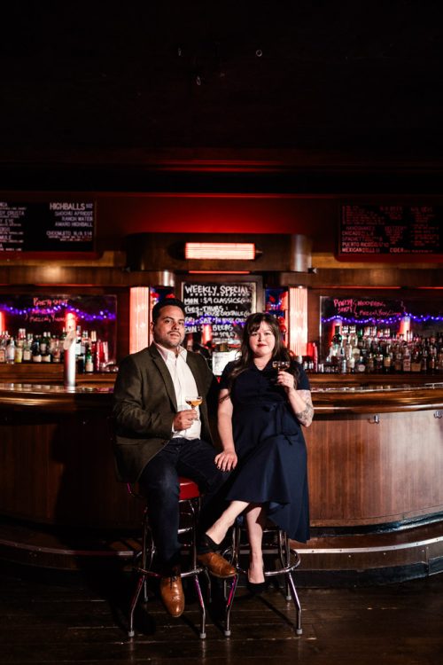 Portrait of engaged couple sitting on bar stools with drinks surrounded by warm red lighting in the California Clipper cocktail bar in Chicago