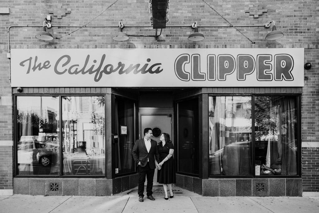 A couple announces their engagement outside The California Clipper, a classic cocktail bar, captured in black and white.