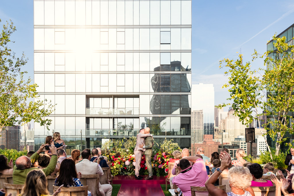 Two grooms kiss on stage surrounded by colorful florals and loved ones at their Chicago rooftop wedding ceremony at 167 Events