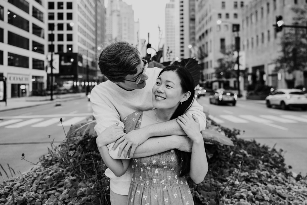 Black and white photo of engaged couple holding each other while standing on a Michigan Avenue median in downtown Chicago