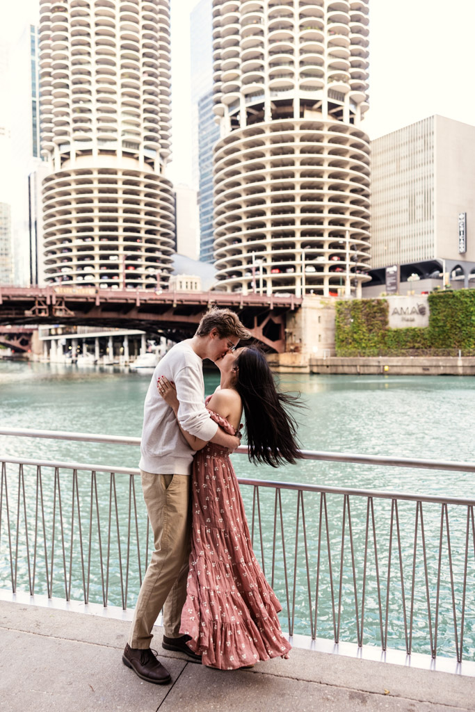 Couple kisses while standing on the Chicago Riverwalk with the Marina Towers visible behind them during downtown engagement session