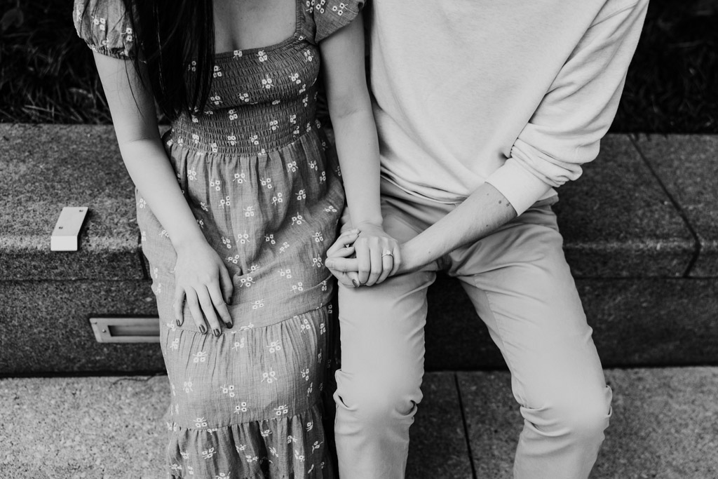 Black and white photo of couple holding hands while sitting on a granite ledge at the Chicago Riverwalk with woman's engagement ring visible