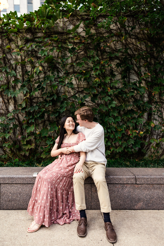 Engaged couple sits on a granite ledge at the Chicago Riverwalk with an ivy wall behind them